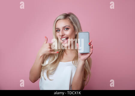 Gerne blonde Frau zeigt der Bildschirm des Smartphones überrascht und zeigen auf, während Sie in die Kamera schaut mit offenen Mund über rosa Hintergrund Stockfoto