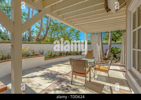 Haus mit einem Außenbereich auf der Terrasse mit weißen Vorhängen. Weißen Stein Zaun, blühende Bäume, und die üppigen Bäume können um den Bereich gesehen werden. Stockfoto