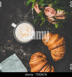 Kaffeepause. Flachbild-lay von Tasse heißen Cappuccino mit frischen Croissants, Blumenstrauß aus rosa Blüten und Bettwäsche Serviette über schwarzen schäbig Hintergrund, Ansicht von oben Stockfoto