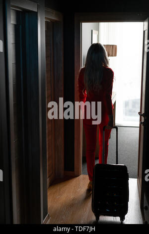 Blonde junge geschäftsfrau kommt in ein Hotelzimmer mit schwarzen Koffer. Frau in Rot coral Business Suit. Junge Mädchen packs Koffer. Stockfoto