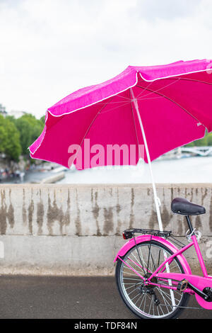 Rosa Fahrrad mit Regenschirm. Fahrrad unter einem rosa Regenschirm auf dem Hintergrund einer konkreten Zaun. Stockfoto