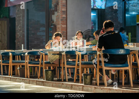 Chengdu, Provinz Sichuan, China - Juni 12, 2019: Junge Chinesen essen außerhalb ein Restaurant in einer Straße am späten Nachmittag Stockfoto