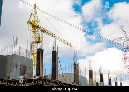 Der Prozess des Aufbaus einer mehrstöckigen Wohnhaus, ein gelber Turm Kran, die konkrete Spalten mit Fittings vor einem blauen Himmel mit Wolken Stockfoto