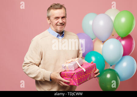 Jolly schöne Reife bärtige Mann in Pullover gegen rosa Hintergrund mit bunten Luftballons und Holding Geburtstag Geschenke Stockfoto