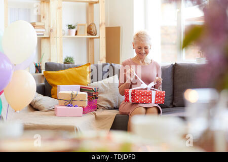Lächelnd aufgeregt reife Lady mit kurzen Haaren sitzen auf komfortablen Sofa und Eröffnung Geschenkbox nach Geburtstagsfeier zu Hause Stockfoto