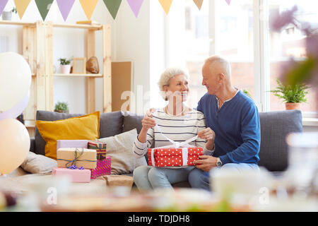 Positive aufgeregt wunderschönen senior Paar in casual Outfits sitzen auf dem Sofa und präsentiert nach der Geburtstagsfeier Stockfoto