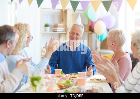 Gruppe von aufgeregt Freunde am Esstisch und applaudieren Geburtstag Mann, während ihn beglückwünschen mit Feier am Abend sitzen Stockfoto
