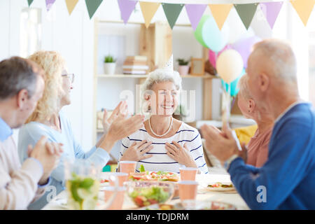 Gruppe der besten Freunde am Esstisch und applaudieren glücklich pensionierte Dame, während ihr zunächst mit dem Geburtstag sitzen Stockfoto