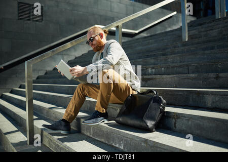 Schwere moderne bärtigen Manager in Sonnenbrille sitzt auf der Treppe und Analyse online Bericht über Tablet Stockfoto