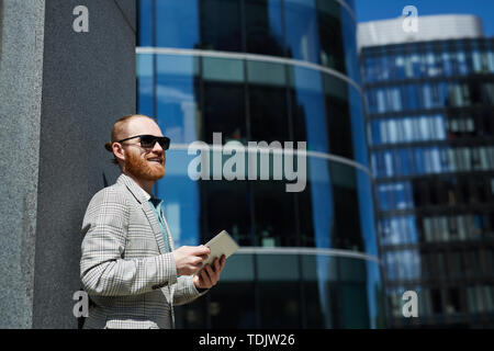 Lächelnde junge unternehmungslustige Manager in hipster Kleidung stehen im Business Viertel und das Arbeiten mit Daten online auf Tablet Stockfoto