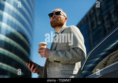 Positiv ehrgeizig stattlichen hipster Geschäftsmann in stilvollen Brillen lehnte sich auf Auto- und trinken Kaffee auf Urban Street Stockfoto
