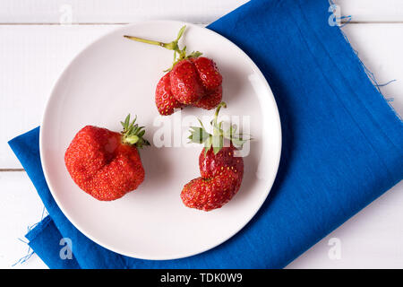 Flach drei Heimische hässlich Erdbeeren auf weiße Platte, blau serviette, über weiß Holz- Hintergrund. Stockfoto