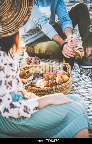 Sommer Picknick am Strand bei Sonnenuntergang. Junges Paar in Wochenende Picknick im Freien am Meer mit frischem Obst und leckeren Häppchen und Öffnen der Flasche Stockfoto
