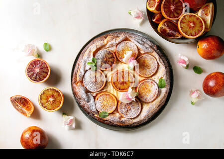 Hausgemachter Käsekuchen mit sizilianischer Blutorangen, dekoriert von essbaren Blüten, Minze und Zucker Pulver in der Platte mit geschnittenen Orangen oben o serviert. Stockfoto