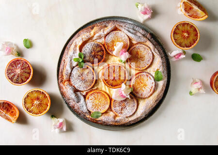 Hausgemachter Käsekuchen mit sizilianischer Blutorangen, dekoriert von essbaren Blüten, Minze und Zucker Pulver in der Platte mit geschnittenen Orangen oben o serviert. Stockfoto