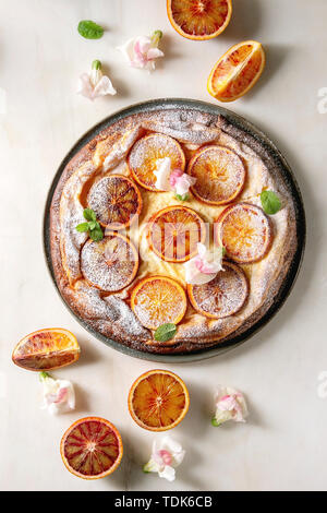 Hausgemachter Käsekuchen mit sizilianischer Blutorangen, dekoriert von essbaren Blüten, Minze und Zucker Pulver in der Platte mit geschnittenen Orangen oben o serviert. Stockfoto