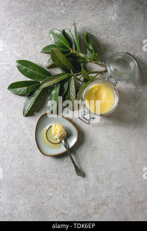 Hausgemachte geschmolzen Ghee geklärte Butter im offenen Glas Glas und Löffel auf Untertasse über grau Textur Hintergrund mit grünen Zweig. Flach, Raum Stockfoto