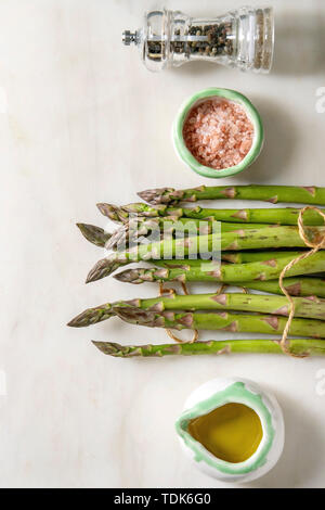 Bündel von organischen grüner Spargel mit Zutaten zum Kochen rosa Meersalz, schwarzer Pfeffer und Olivenöl in weißem Marmor Hintergrund. Flach, Raum Stockfoto