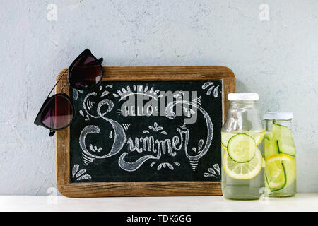 Sommer Thema. Chalk Schriftzug Hallo Sommer auf vintage Tafel, Sonnenbrille und zwei Glasflaschen mit sassy Zitrone und Gurken Wasser auf weißem Marmor Stockfoto