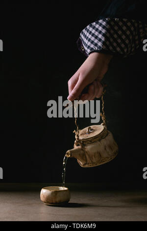Die Hand des jungen Mannes gießen aus handgefertigtem Handwerk Teekanne heiße grüner Tee in der traditionellen chinesischen Ton Keramik Schale stehend auf dunklen Tisch mit schwarzem Hintergrund Stockfoto