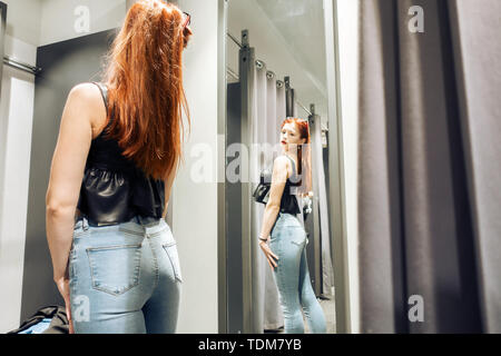 Junge schlanke Mädchen, das versuchte auf neuen Jeans im Proberaum im Store. Frau mit schönen Abbildung sieht sich selbst in der Reflexion der Spiegel. Pu Stockfoto