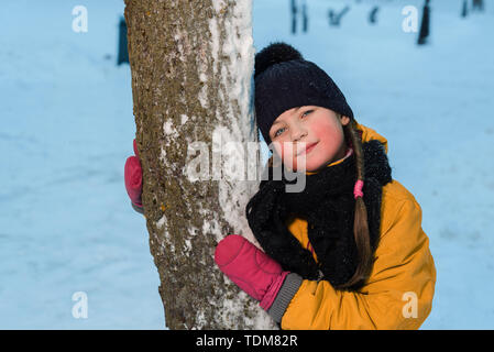 Winter Portrait von ein nettes kleines Mädchen in der Nähe von einem Baum. Das Kind trägt ein Winter Mütze und Handschuhe Stockfoto