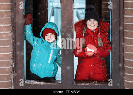 Glückliche Kinder. Bruder und Schwester spielen Schneebälle im Winter. die Kinder sind ein Lächeln auf den Lippen und Suchen aus den Fenstern der ein Holzhaus.boy seine Hand winken Stockfoto