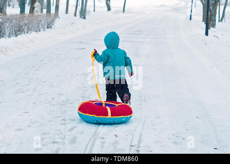 Ein kleiner Junge im Winter verläuft entlang der schneebedeckten Straße allein. Kind zieht Schlitten Schläuche Stockfoto