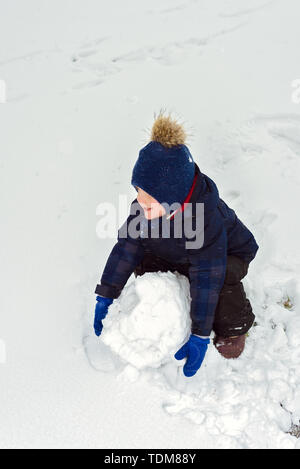 Kleiner Junge macht ein Schneemann im Winter. Kind rollt einen Schneeball. glückliches Kind spielt und lächelt. Emotion und Glück auf Ferien für Kinder Stockfoto