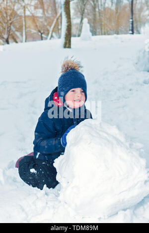 Kleiner Junge macht ein Schneemann im Winter. Kind rollt einen Schneeball. glückliches Kind spielt und lächelt. Emotion und Glück auf Ferien für Kinder Stockfoto