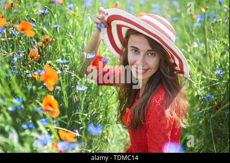 Verträumte Frau im roten Kleid und einem grossen Rot gestreifte Hut sitzt im schönen Kraut blühenden Mohnfeld Sommer. Vintage elegant romantisch aussehen. Konzept Stockfoto
