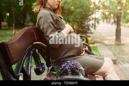 Schwangere Frau sitzt auf der Bank im Park, in der Nähe von ihrem Bauch. Grüne Bäume und Gasse Hintergrund. Stockfoto