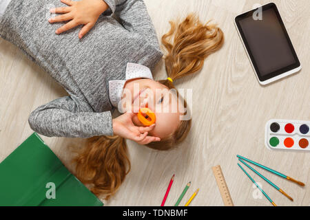 Süße kleine Mädchen mit O Buchstaben auf dem Boden liegt. Das Kind ist durch ein Buch, Tablet, Farben, Pinsel, Stifte umgeben. Stockfoto