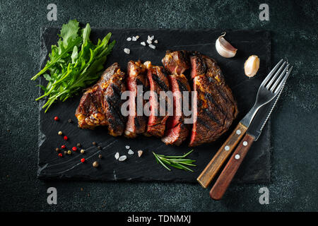 Ribeye Steak in Scheiben, gegrillt mit Pfeffer, Knoblauch, Salz und Thymian auf einer Schiefertafel Schneidebrett auf einem dunklen Stein serviert. Ansicht von oben. Flach. Stockfoto