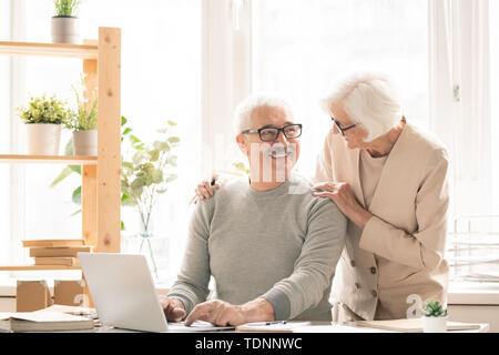 Gerne älterer Mann in der casualwear sitzen Schreibtisch vor Laptop, während im Gespräch mit seiner Frau neben ihm stehen Stockfoto