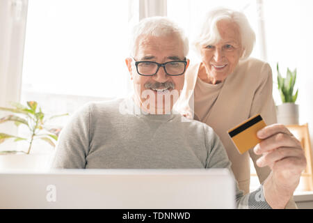 Zeitgenössische älterer Mann mit Plastikkarte sitzen vor dem Laptop online Einkaufen und Bezahlen für Aufträge Stockfoto