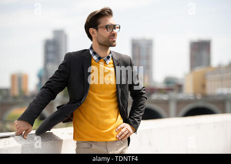 Elegante junge Unternehmer in Jacke, gelb Pullover und Jeans ständigen von Riverside in städtische Umwelt Stockfoto