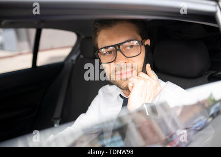 Zuversichtlich, dass junge Unternehmer in Brillen im Auto sitzen, mit dem Auto zur Arbeit fahren und im Gespräch mit Kunden auf dem Smartphone Stockfoto