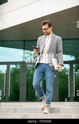 Junge schwere beschäftigter Manager in Smart Casual Bewegen unten beim Blättern im Smartphone nach der Arbeit Stockfoto
