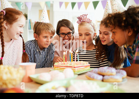 Portrait von glücklichen kleinen Mädchen bläst die Kerzen auf der Geburtstagstorte während der Party mit Freunden, kopieren Raum Stockfoto