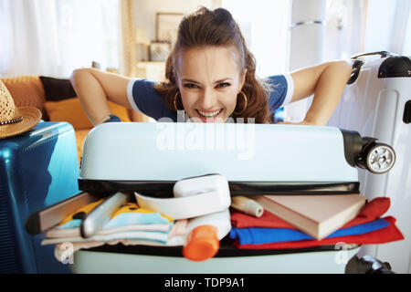 Lächelnd elegante reisenden Frau in Blau t-shirt im modernen Haus im sonnigen Sommertag versuchen, Überlast Tasche zu schliessen. Stockfoto