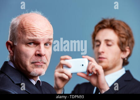 Charismatische konservativen älteren Geschäftsmann ziehen eine wry fragende Gesicht in die Kamera als ein jüngerer Kollege oder Partner zu fotografieren Ihn bereitet auf Stockfoto