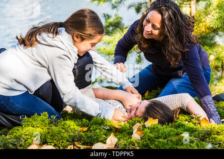 Eine Mutter und ihre zwei Töchter durchgeklickt und Kitzeln einander an einem See bei einem Familienausflug in einem Stadtpark an einem warmen Herbst Tag Stockfoto