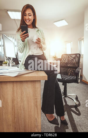 Eine erfolgreiche weibliche Unternehmer sitzt im Büro Tisch und Schreiben von Nachrichten auf einem Smartphone und Getränke Kaffee im Büro. Stockfoto