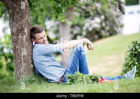 Mit frischem Gemüse auf dem Rasen im Garten liegen, lehnte sich gegen einen Baumstamm, Deutschland Stockfoto