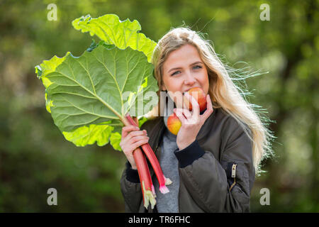 Rhabarber (Rheum rhabarbarum), junge blonde Frau mit frisch gepflückt Rhabarber mit Vergnügen in einen Apfel beissen, Deutschland Stockfoto