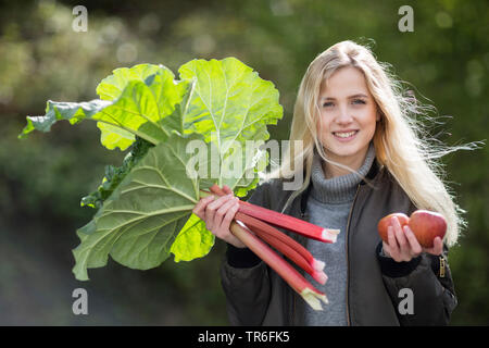 Rhabarber (Rheum rhabarbarum), junge blonde Frau mit frisch gepflückt Rhabarber und Äpfel in den Händen, Deutschland Stockfoto