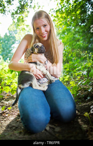 Haushund (Canis lupus f. familiaris), gut aussehende junge Frau mit einem puggle im Arm, kniend auf Waldboden, Deutschland Stockfoto