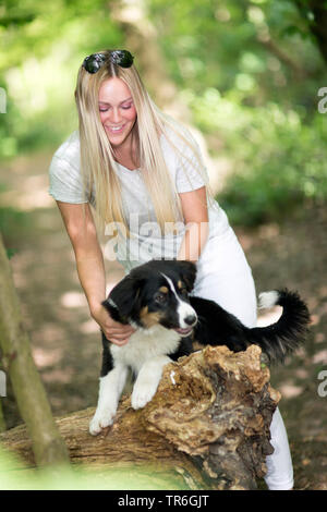 Australian Shepherd (Canis lupus f. familiaris), Welpe mit einer jungen Frau auf einem Waldweg an einer toten Baumstamm, Deutschland Stockfoto