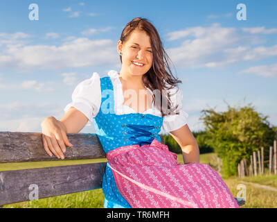 Junge Frau im Dirndl auf einer Bank sitzen auf einer Bergwiese, Deutschland, Bayern Stockfoto